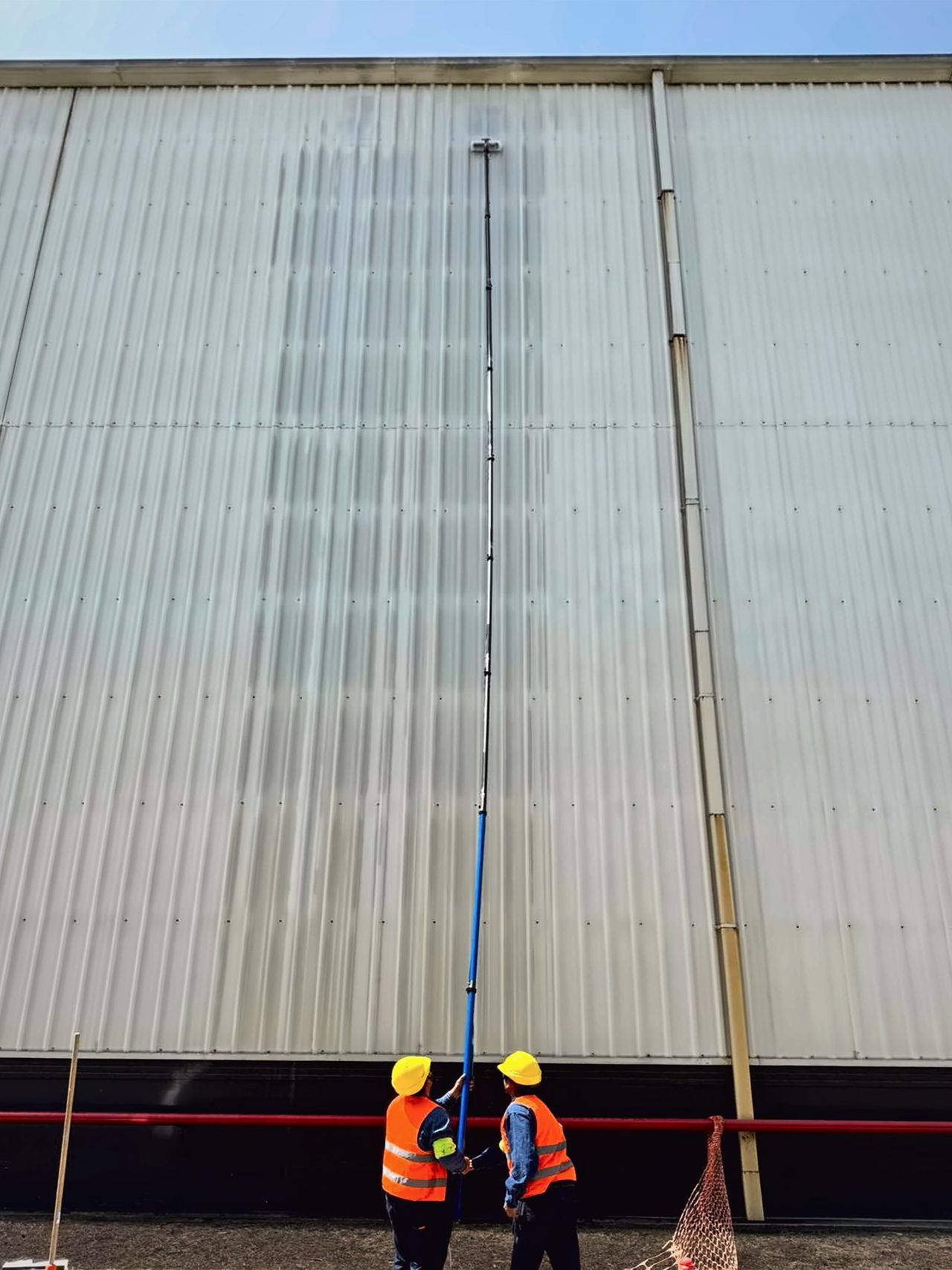 Dos operarios con casco y chaleco naranja limpian desde el piso la fachada de una nave industrial con una pértiga telescópica de fibra de carbono, bajo cielo azul.