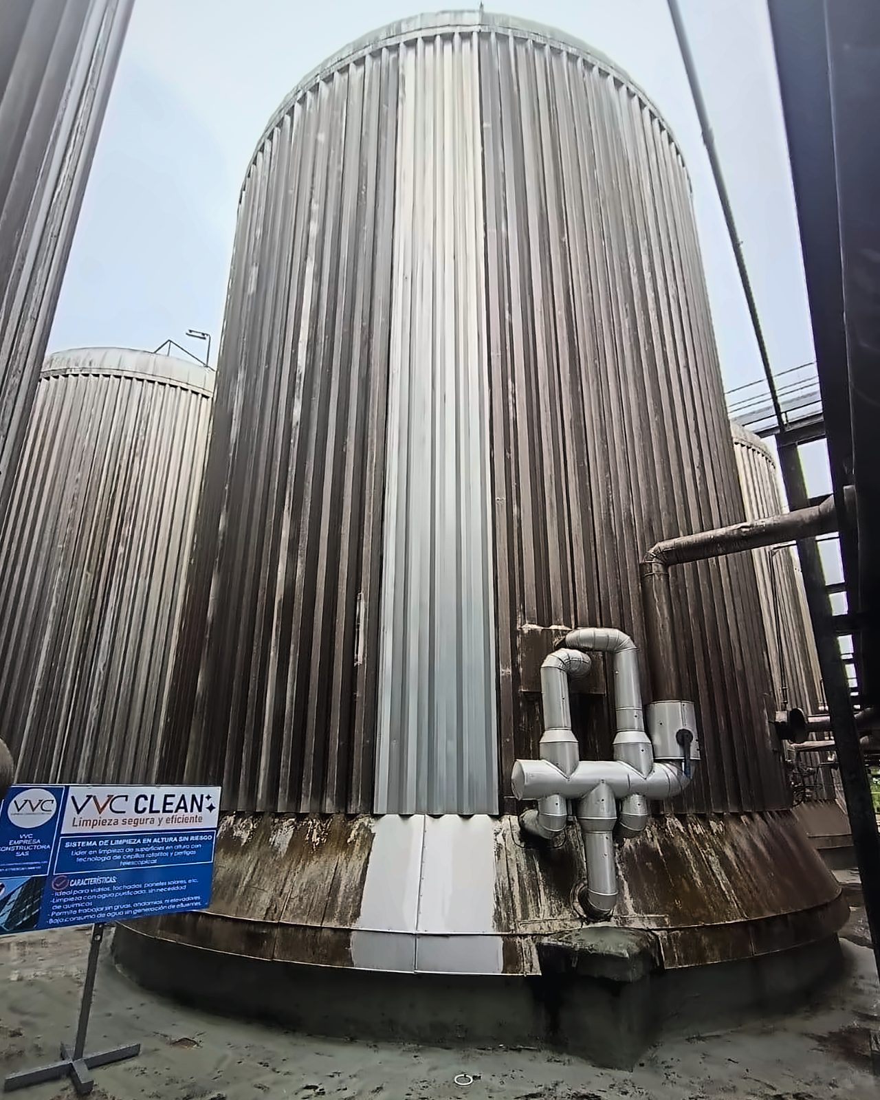 Tanque industrial de acero corrugado con una franja vertical recién limpiada por VVC Clean: el antes y el después en una sola toma, con el cartel de la empresa abajo a la izquierda.